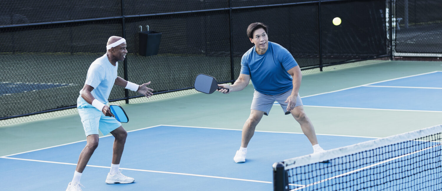 Two multi-ethnic senior men playing pickleball. They are a doubles team. The African-American man, in his 70s, is watching as his partner, an Asian man in his 60s, gets ready to hit the ball with his paddle.